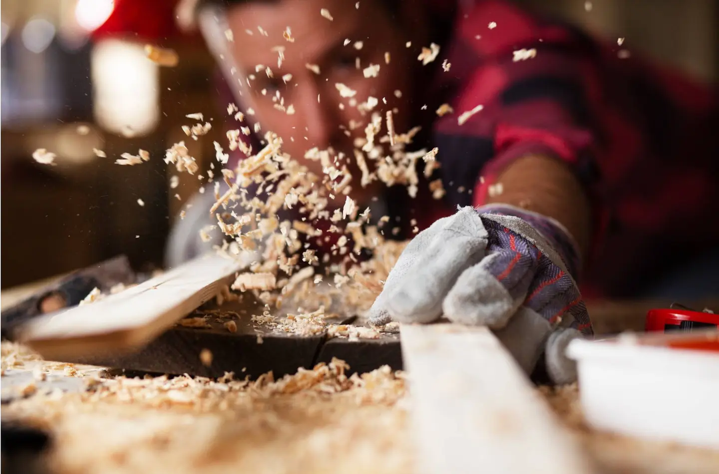 Close-up van een persoon die een houten plank schuurt, terwijl houtkrullen rondvliegen. De persoon draagt handschoenen en een rood-zwart geruit overhemd.