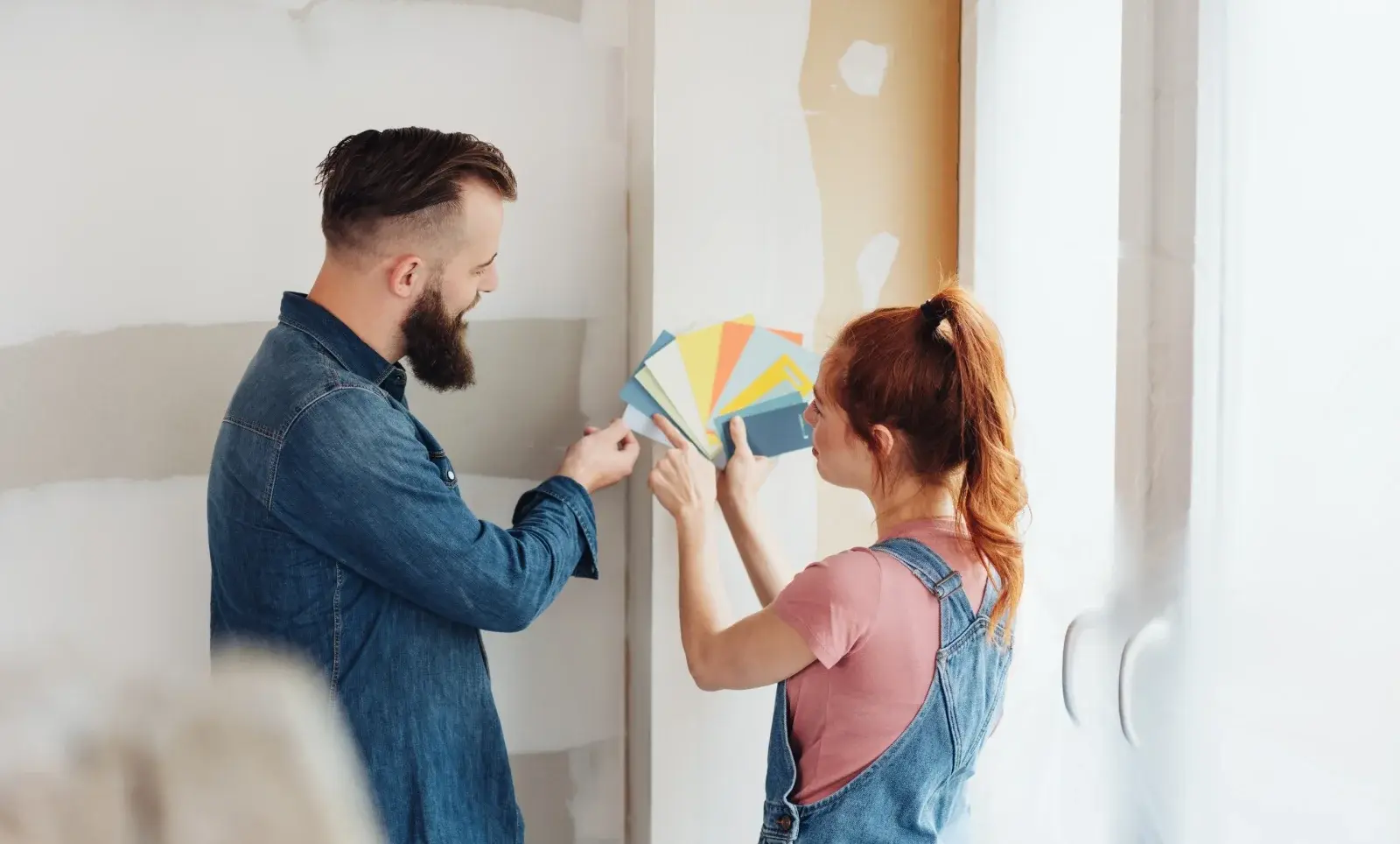 Een man met een baard in een spijkerblouse en een vrouw met rood haar in een overall en een T-shirt houden verfstalen vast en bespreken de kleurkeuze voor de muur in een onafgemaakte kamer. Ze lijken opgewonden als ze nadenken over Viva Magenta, de Kleur van het Jaar 2024.