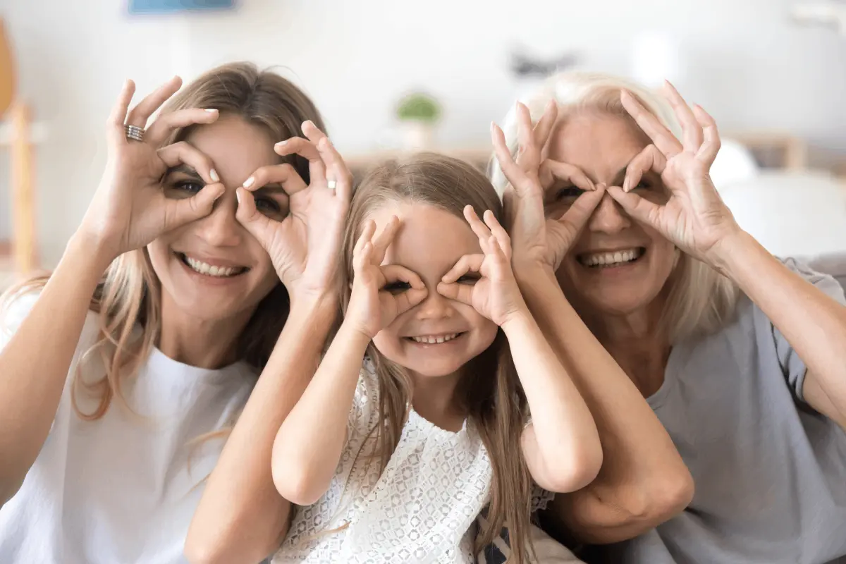 Drie mensen, een volwassen vrouw, een kind en een oudere vrouw, zitten naast elkaar en maken een speelse bril met hun handen voor hun ogen.