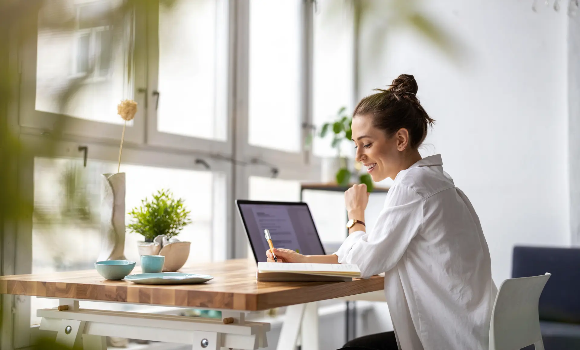 Een persoon werkt aan een bureau met een laptop en een notitieboekje, met een vaas, planten en een koffiekopje op het bureau, naast grote ramen.