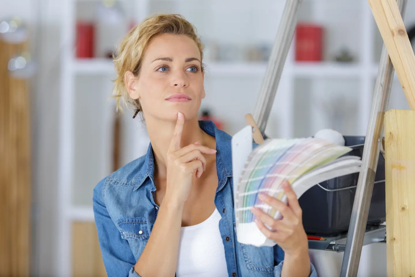 Show thumbnail preview Een vrouw met kort blond haar, gekleed in een blauw spijkerblouse, houdt een boekje met Pantone-kleurenstalen vast en kijkt peinzend opzij. Ze staat naast een ladder in een goed verlichte kamer