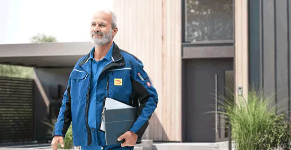 Een man in een blauw jasje met documenten in zijn hand staat voor een modern gebouw met een houten buitenkant.