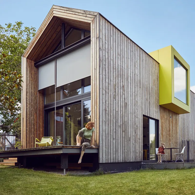 Modern houten huis met grote ramen en een felgele accentdoos. Een persoon zit op de rand van de veranda en een andere persoon zit vlakbij. Het huis is omgeven door groen.