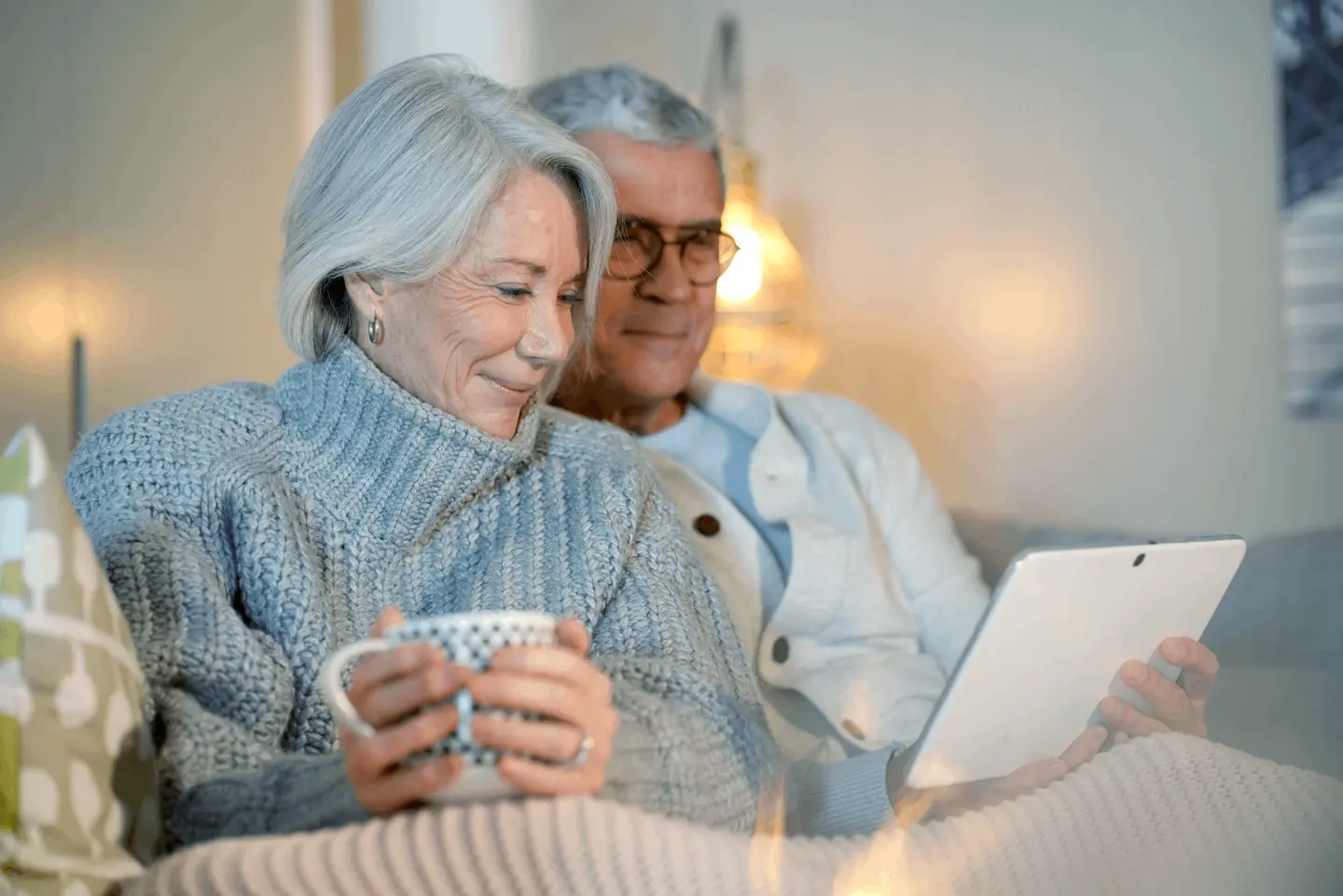 Een ouder echtpaar zit comfortabel samen op een bank, de een houdt een mok met patroon vast en de ander een tablet. Beiden glimlachen en lijken gefocust op het tabletscherm.