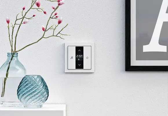 Een moderne digitale thermostaat is op een witte muur gemonteerd naast een ingelijste foto. Op een plank eronder staan twee decoratieve glazen vazen en een tak met roze bloemen.