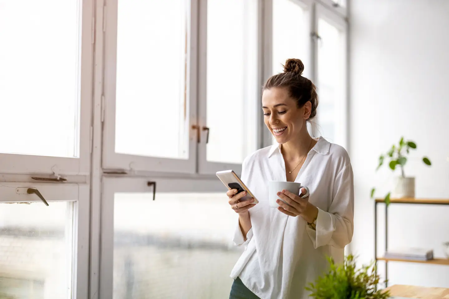 Een vrouw staat glimlachend voor grote ramen en houdt een smartphone in de ene hand en een mok in de andere.